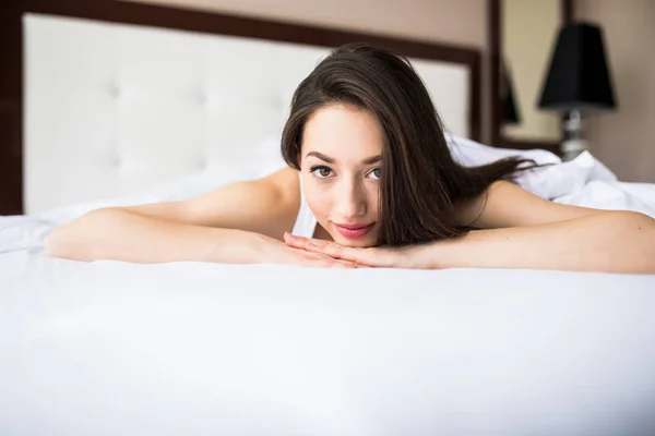 Mujer acostada al final de la cama debajo de la colcha y sonriendo, con la cabeza apoyada sobre la mano con la otra en el pelo . —  Fotos de Stock