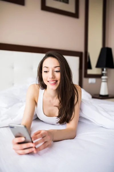 Красивая молодая женщина улыбается и держит смартфон, лежа в постели дома — стоковое фото