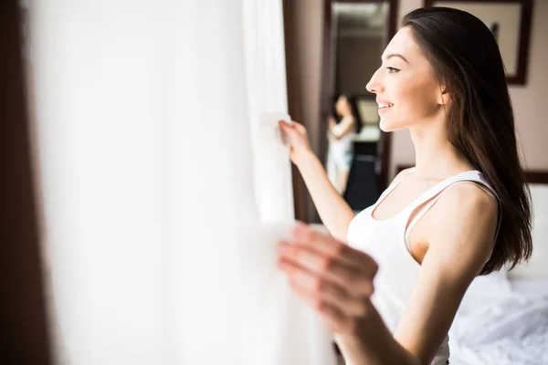 Fiatal nő megnyitja függönyök nézni a kilátás egy ablak reggel — Stock Fotó