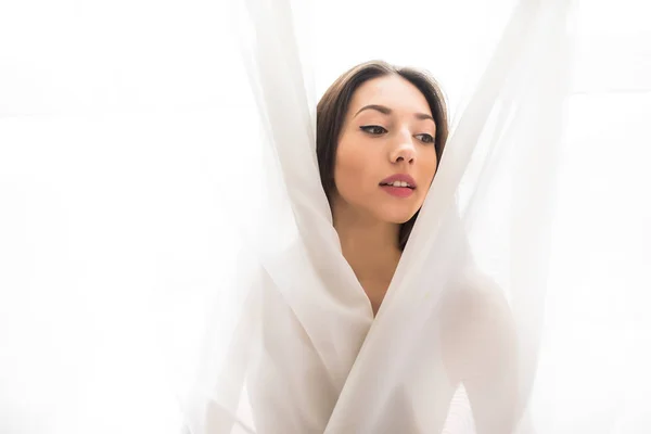 Bella giovane donna guardando da dietro una tenda bianca a casa al mattino, umore felice — Foto Stock