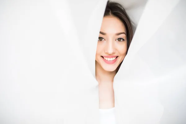 Hermosa mujer joven mirando desde detrás de una cortina blanca en casa por la mañana, estado de ánimo feliz — Foto de Stock