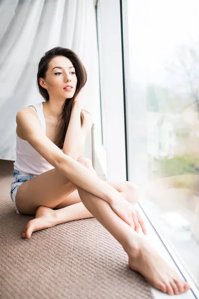 Mujer joven sentada en el suelo abrazando sus rodillas, mirando a través de una ventana de cuerpo entero a la gran ciudad . — Foto de Stock