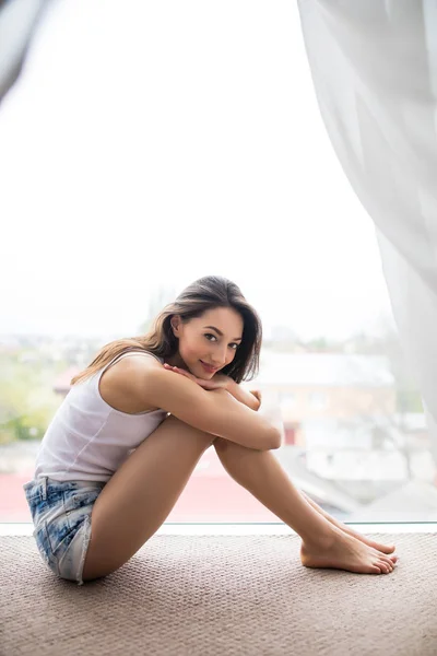 Junge Frau sitzt auf der Fensterbank und schaut morgens zu Hause auf die Stadt — Stockfoto