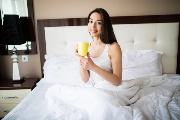 Hermosa mujer bebiendo un café sentada en su cama en casa por la mañana —  Fotos de Stock