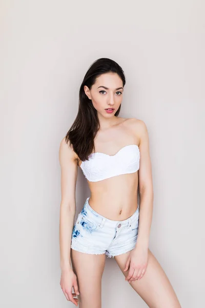 Beautiful young woman with a sports figure in denim shorts and a black bra standing on a white background — Stock Photo, Image