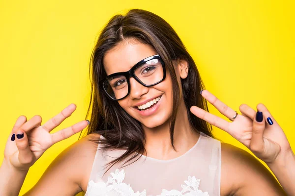 Mladá dívka, která nosí brýle s gestem vítězství v žlutém pozadí. Dívka na kameru. — Stock fotografie
