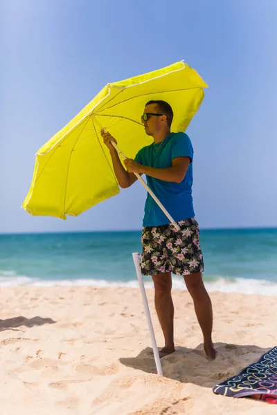 Молодой человек устанавливает в песок солнечный зонтик на пляже возле океана — стоковое фото