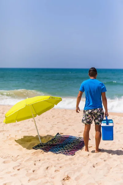 Молодой человек с холодильником для мороженого под солнечным зонтиком на пляже — стоковое фото
