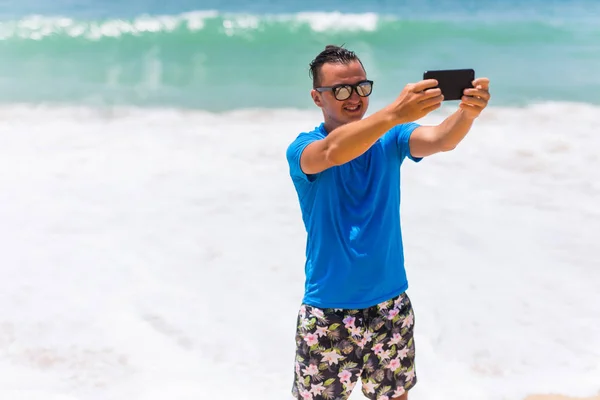Счастливый красивый улыбающийся мужчина в отпуске смеется на пляже, делая селфи — стоковое фото