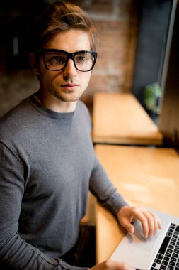 Genç adam bir fincan kahve tutan bir pencereden bakarak bir dizüstü bilgisayar ile 