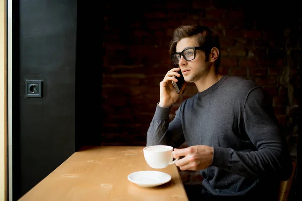 朝はコーヒーを飲みながら窓際で電話で話しているハンサムな若い男 — ストック写真