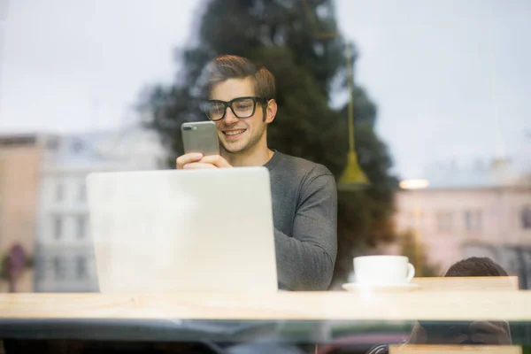 Joven hombre guapo freelancer escribir sms o utilizar el teléfono en frente de la vista del ordenador portátil desde la ventana — Foto de Stock