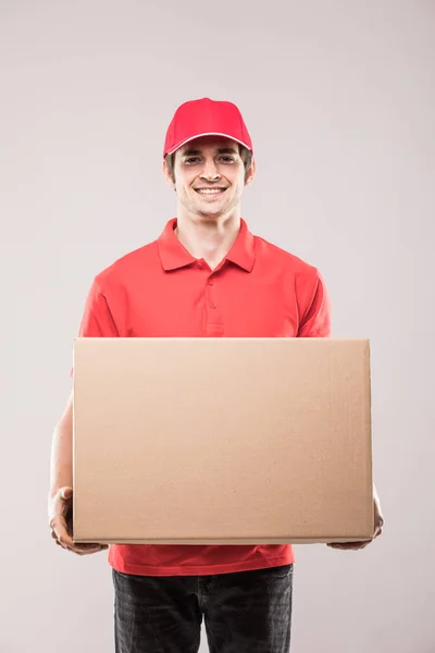 Joven repartidor sonriente con caja en la mano en uniforme rojo sobre blanco — Foto de Stock
