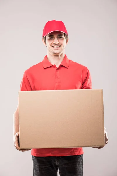 Joven repartidor sonriente con caja en la mano en uniforme rojo sobre blanco — Foto de Stock