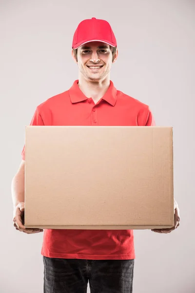 Alegre repartidor feliz joven mensajero sosteniendo una caja de cartón y sonriendo — Foto de Stock