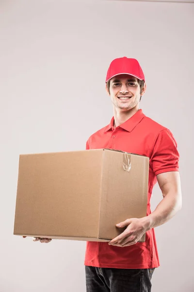 Alegre repartidor feliz joven mensajero sosteniendo una caja de cartón y sonriendo mientras está de pie en gris —  Fotos de Stock