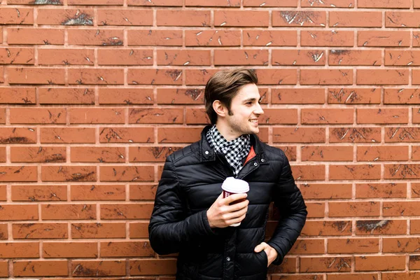 Genç bir adam elinde kahvesiyle bir tuğla duvar zemin karşı — Stok fotoğraf