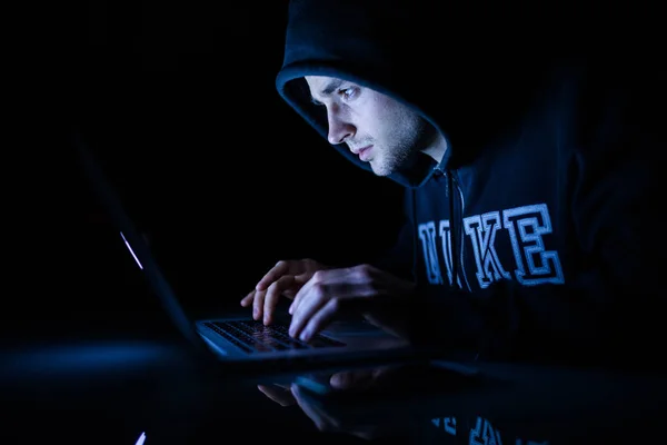 Man in a hood working in front of a laptop hacker cracking programs in the night — Stock Photo, Image