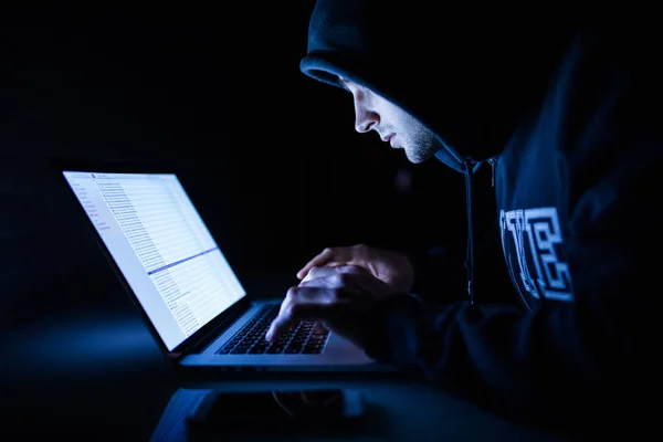 Man in a hood working in front of a laptop hacker cracking programs in the night — Stock Photo, Image