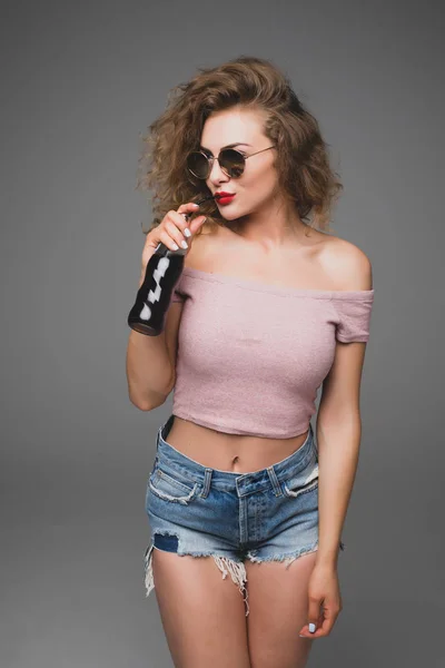 Carino elegante giovane donna riccia bere soda dalla bottiglia su sfondo grigio — Foto Stock