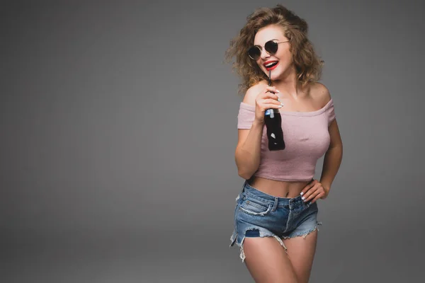 Roztomilý stylový kudrnaté mladá žena pít sodovku z láhve nad šedým pozadím — Stock fotografie