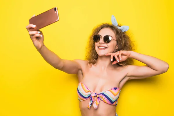 Jeune belle femme bouclée avec sourire heureux dans les lunettes de soleil prendre selfie sur le téléphone sur fond jaune — Photo