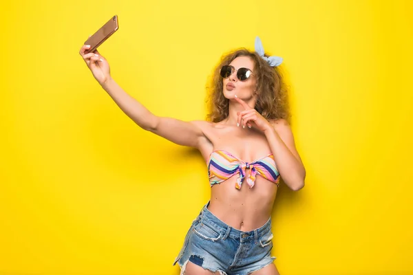 Genç güzel kıvırcık kadın güneş gözlüğü mutlu gülümseme ile telefon Sarı zemin üzerine selfie almak — Stok fotoğraf