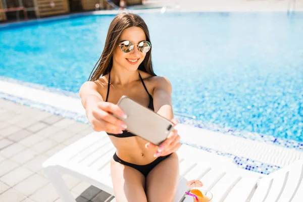 Молодая женщина в купальнике расслабляется с коктейлем у бассейна и делает селфи по телефону — стоковое фото