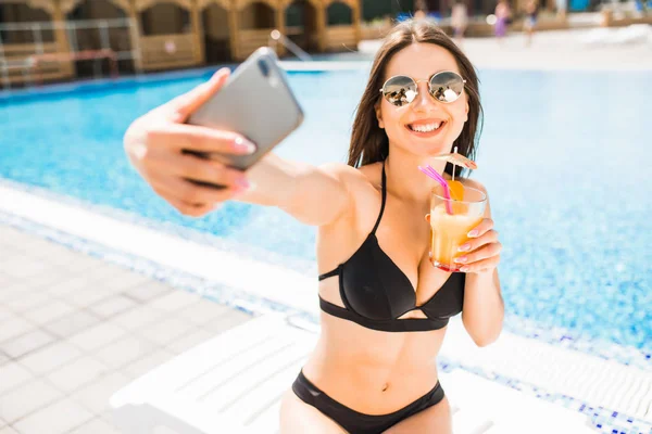 Молодая женщина в купальнике расслабляется с коктейлем у бассейна и делает селфи по телефону — стоковое фото