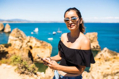 Okyanus kaya üstünde akıllı telefon kullanan Latin güzel kadın. seyahat ve aktif yaşam tarzı kavramı 