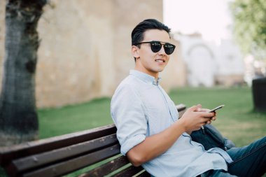 Asya yakışıklı parkta bir bankta cep telefonu kullanarak 