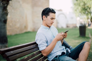 Oturan genç Asyalı adam parkta Bank ile telefon elinde telefon bak.