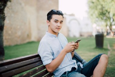 Oturan genç Asyalı adam parkta tezgah onun elinde telefonla kameraya bak.