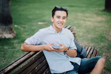 Elleri ve gülümsemeye fotoğraf makinesi telefon ile parkta bankta oturan genç Asyalı adam