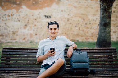 Bir parkta bir bankta oturmuş bir akıllı telefon kullanan mutlu Asyalı adam