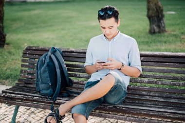 Şehir yakışıklı öğrenci bankta oturmuş akıllı telefon kullanan rahat genç Asyalı adam