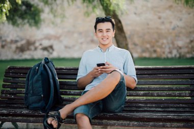 Bir parkta bir bankta oturmuş bir akıllı telefon kullanan mutlu Asyalı adam