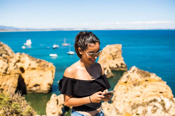 Mulher bonita latina usando telefone inteligente no topo da rocha oceânica. viagem e conceito de estilo de vida ativo — Fotografia de Stock