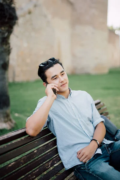 Πορτρέτο της μικρά Ασίας όμορφος άνδρας μιλάει στο τηλέφωνο κάθεται σε παγκάκι σε εξωτερικούς χώρους — Φωτογραφία Αρχείου
