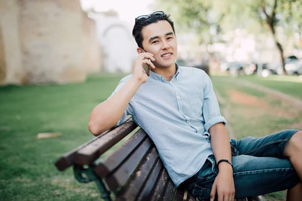 Όμορφος Ασίας άντρας κάθεται στον πάγκο και να μιλάει στο τηλέφωνο σε εξωτερικούς χώρους — Φωτογραφία Αρχείου