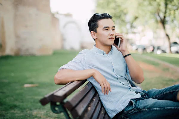 Όμορφος Ασίας άντρας κάθεται στον πάγκο και να μιλάει στο τηλέφωνο σε εξωτερικούς χώρους — Φωτογραφία Αρχείου