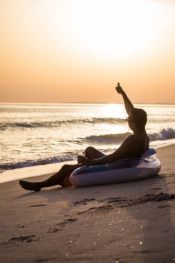 Bir bardak bira float daire, kauçuk disk plaj ve başparmak yukarıya ile hoş geldiniz günbatımı üzerinde oturan genç adam. Yaz meslek