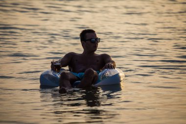 Bir bardak bira Kauçuk halka okyanus su üzerinde yüzen günbatımında güneş gözlüğü genç adam. Yaz meslek.