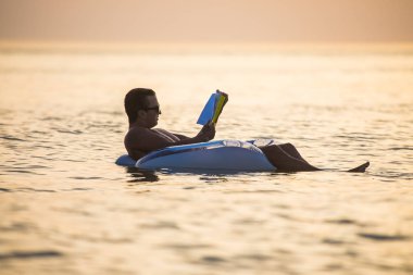 Beyaz adam on sunset okyanus su içerisinde yüzen bir kitap okur. Yaz meslek.