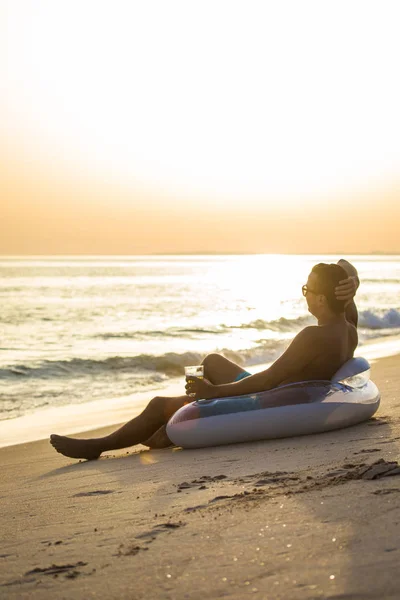 Unge mann med et glass øl på fløtesirkelen, gummiskive på stranden og velkommen solnedgang med hendene over hodet. Sommeryrke – stockfoto