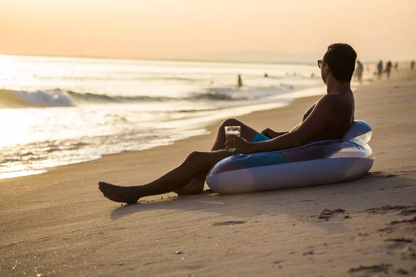 Bir bardak bira float Kauçuk disk plaj ve hoş geldiniz günbatımı üzerinde oturan genç adam. Yaz meslek