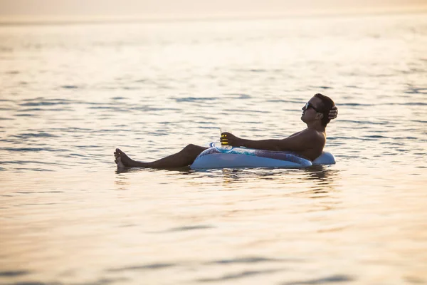 Молодой человек со стаканом пива лежит на резиновом диске в воде на морском закате — стоковое фото