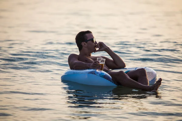 Hombre joven en gafas de sol con un vaso de cerveza flotando en el anillo de goma en el agua del océano al atardecer. Vocación de verano . — Foto de Stock