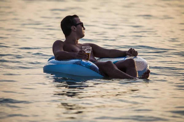 Jonge man in zonnebril kunt u ontspannen in het water van de Oceaan op opblaasbare ring op zonsondergang en drinken bier. Zomer roeping — Stockfoto