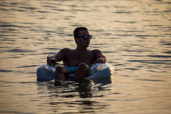 Bir bardak bira Kauçuk halka okyanus su üzerinde yüzen günbatımında güneş gözlüğü genç adam. Yaz meslek.